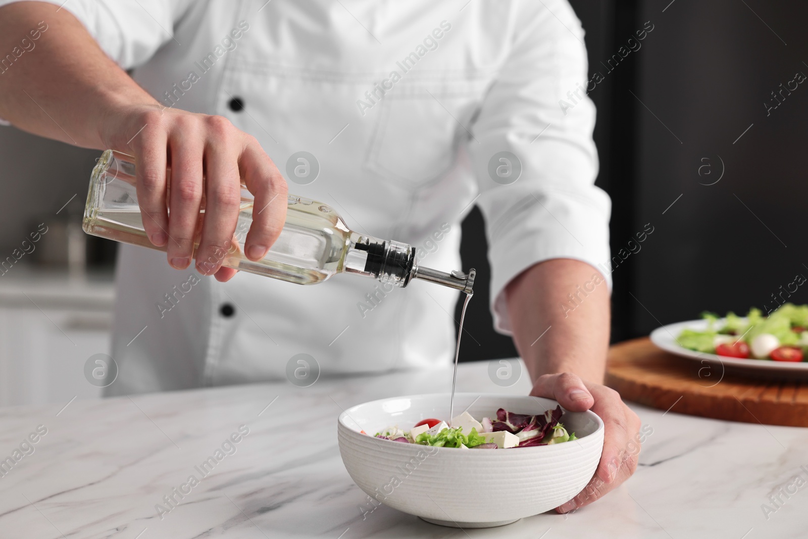 Photo of Professional chef pouring oil onto delicious salad at white marble table in kitchen, closeup