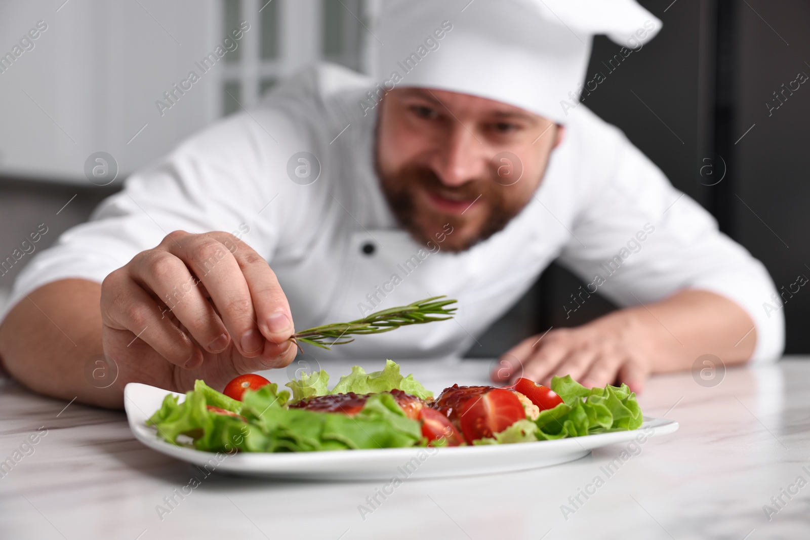 Photo of Professional chef decorating delicious dish with rosemary at white marble table in kitchen, selective focus