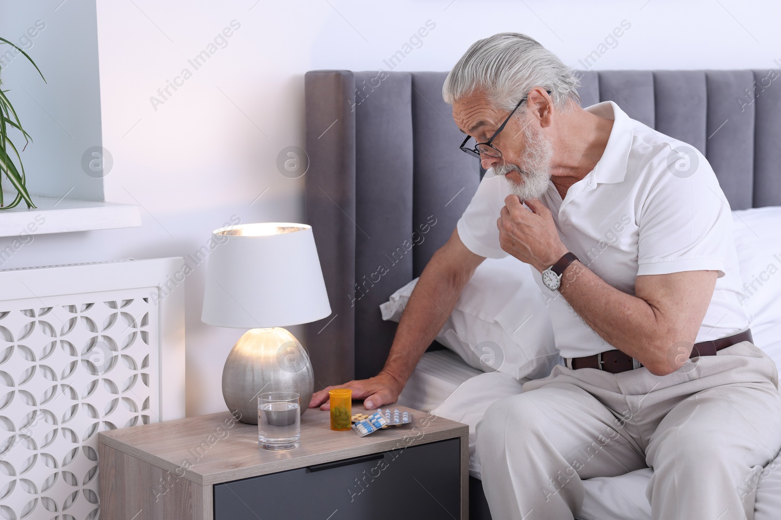 Photo of Senior man near bedside table with pills and glass of water indoors