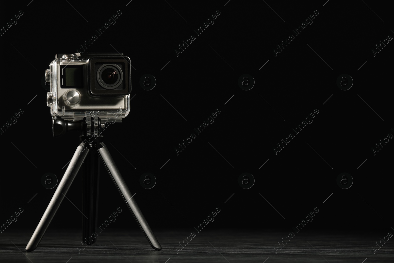 Photo of Modern action camera and tripod on wooden surface against black background. Space for text