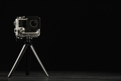 Modern action camera and tripod on wooden surface against black background. Space for text