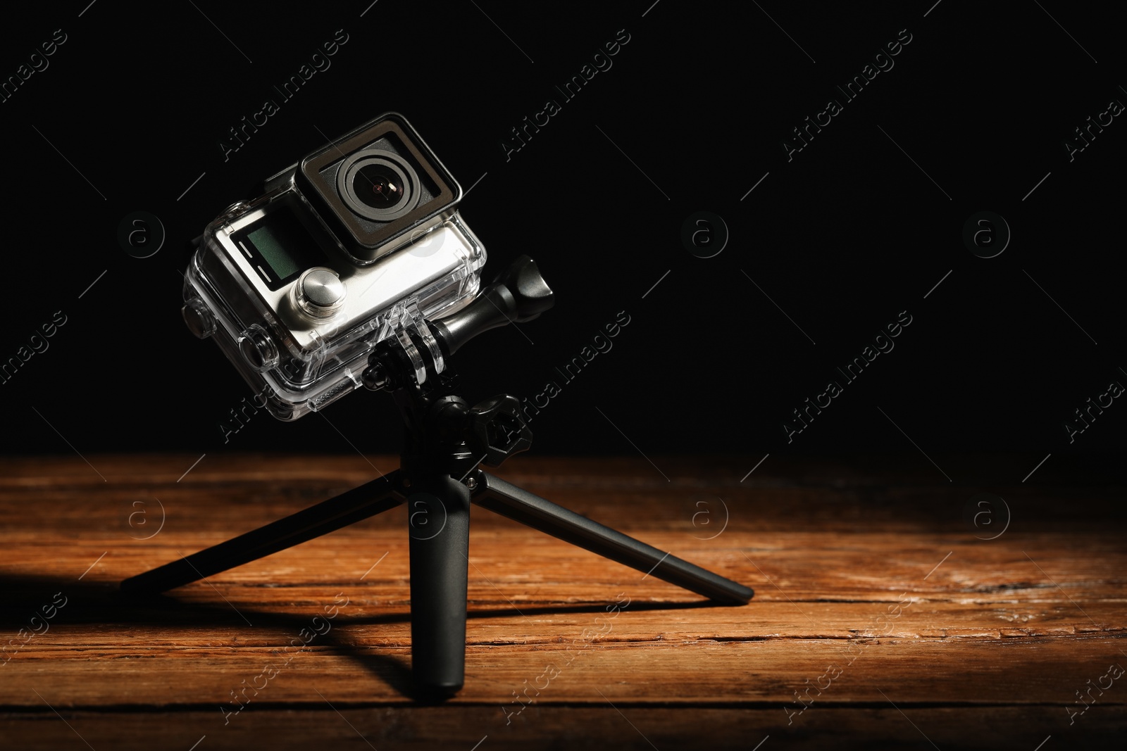 Photo of Modern action camera and tripod on wooden surface against black background. Space for text