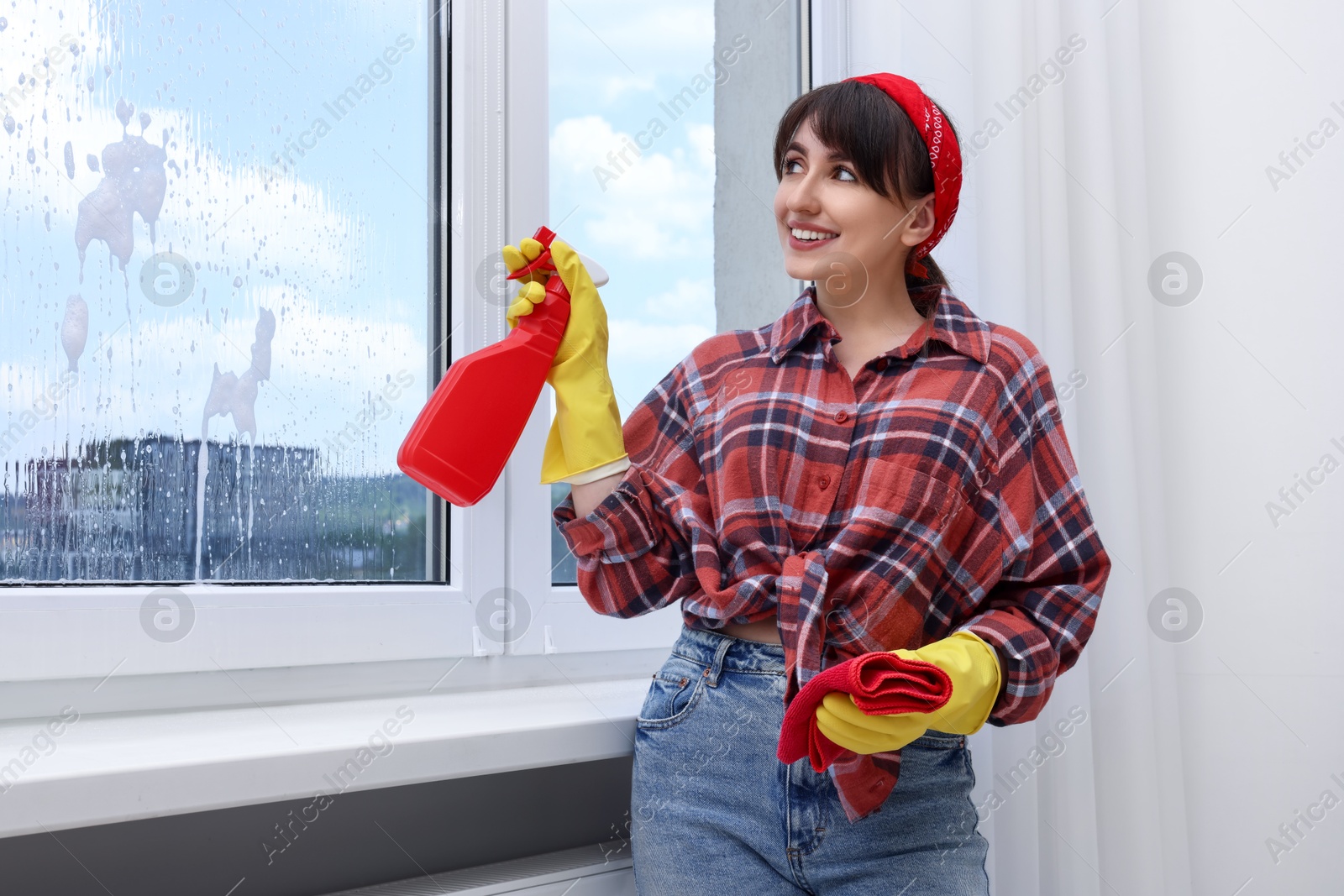 Photo of Beautiful young woman with spray bottle of detergent and napkin cleaning window indoors