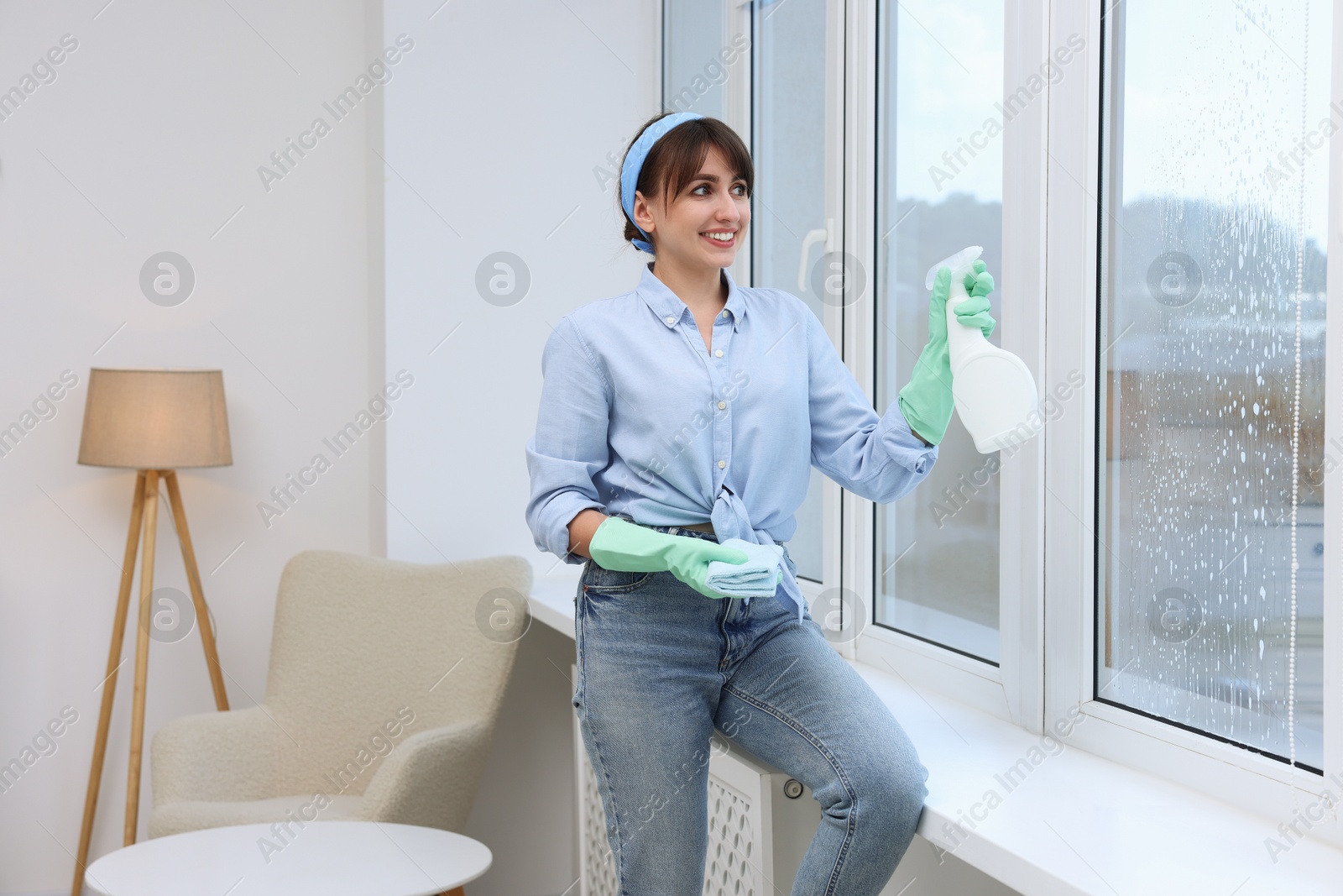 Photo of Beautiful young woman with spray bottle of detergent and napkin cleaning window indoors