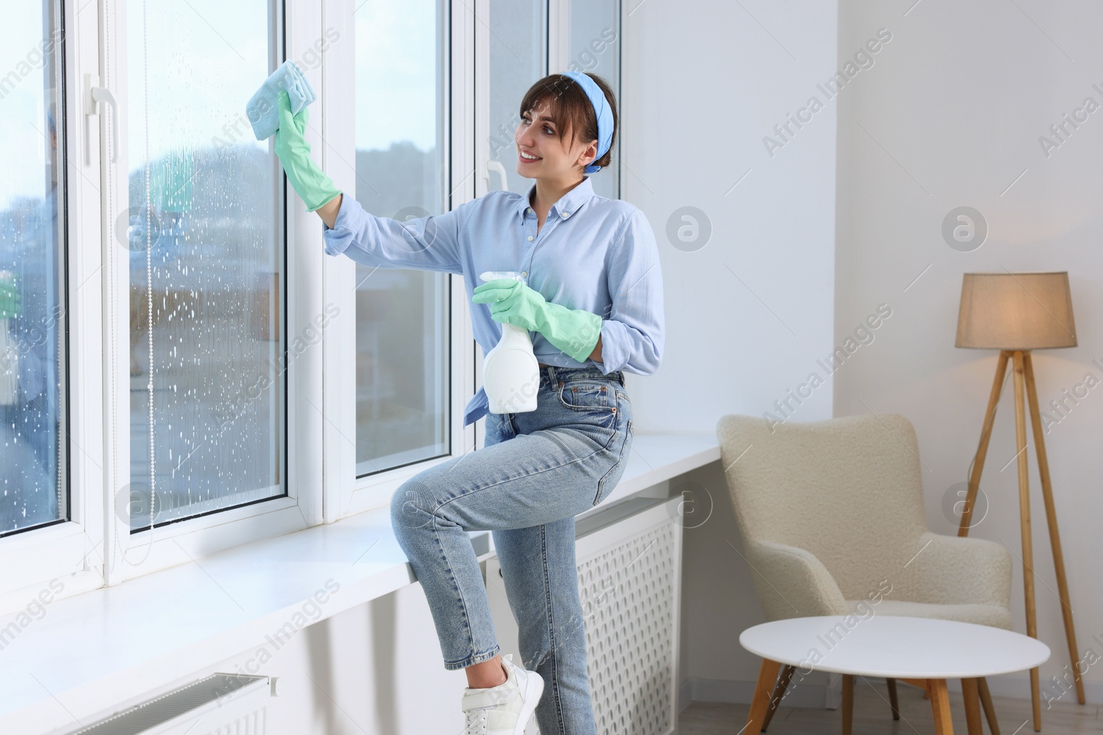 Photo of Beautiful young woman with spray bottle of detergent and napkin cleaning window indoors