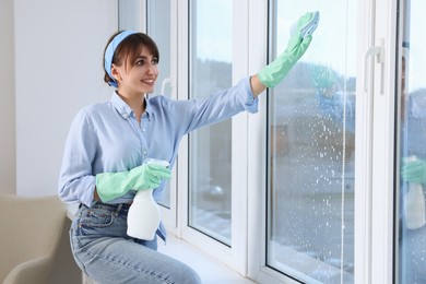 Beautiful young woman with spray bottle of detergent and napkin cleaning window indoors