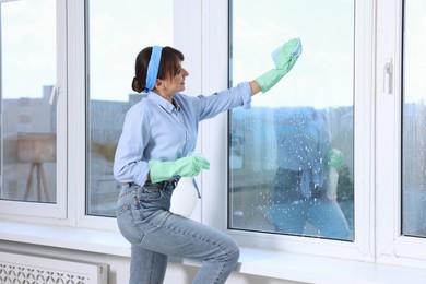 Photo of Beautiful young woman with spray bottle of detergent and napkin cleaning window indoors