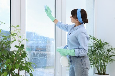 Beautiful young woman with spray bottle of detergent and napkin cleaning window indoors