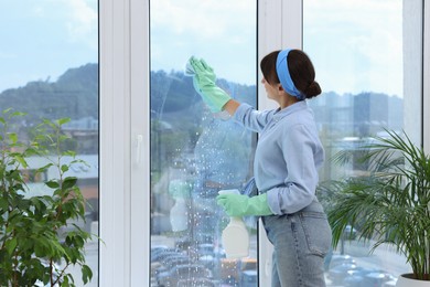 Photo of Young woman with spray bottle of detergent and napkin cleaning window indoors