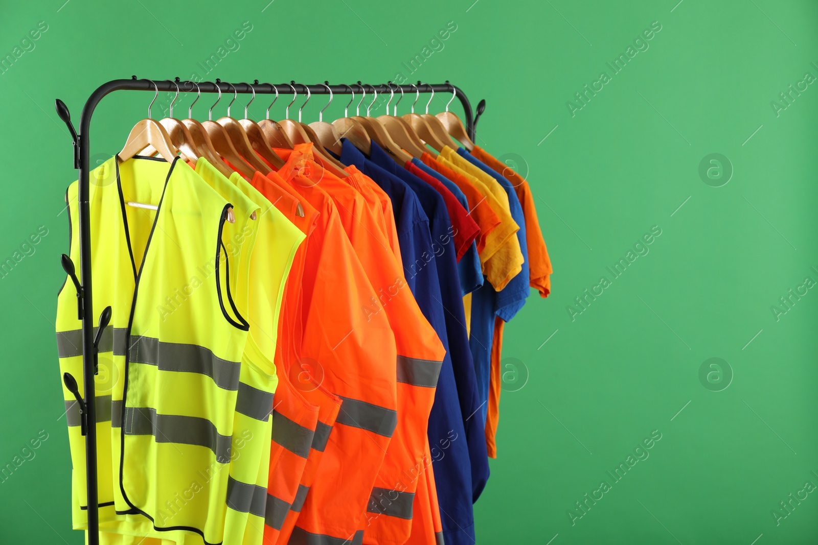 Photo of Different workers' uniforms and reflective vests on clothing rack against green background. Space for text