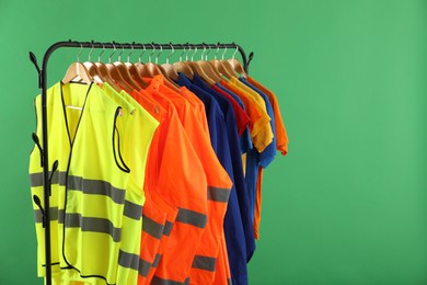 Different workers' uniforms and reflective vests on clothing rack against green background. Space for text