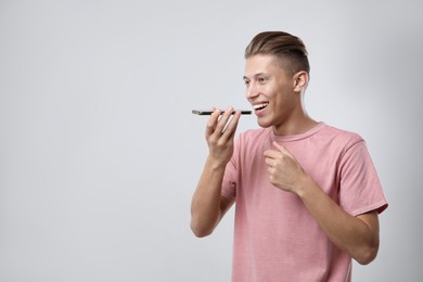 Young man recording voice message via smartphone on light gray background, space for text