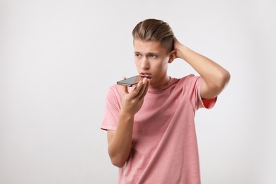 Young man recording voice message via smartphone on light gray background