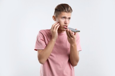 Young man recording voice message via smartphone on light gray background