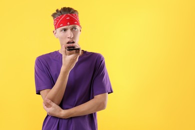 Young man recording voice message via smartphone on yellow background, space for text