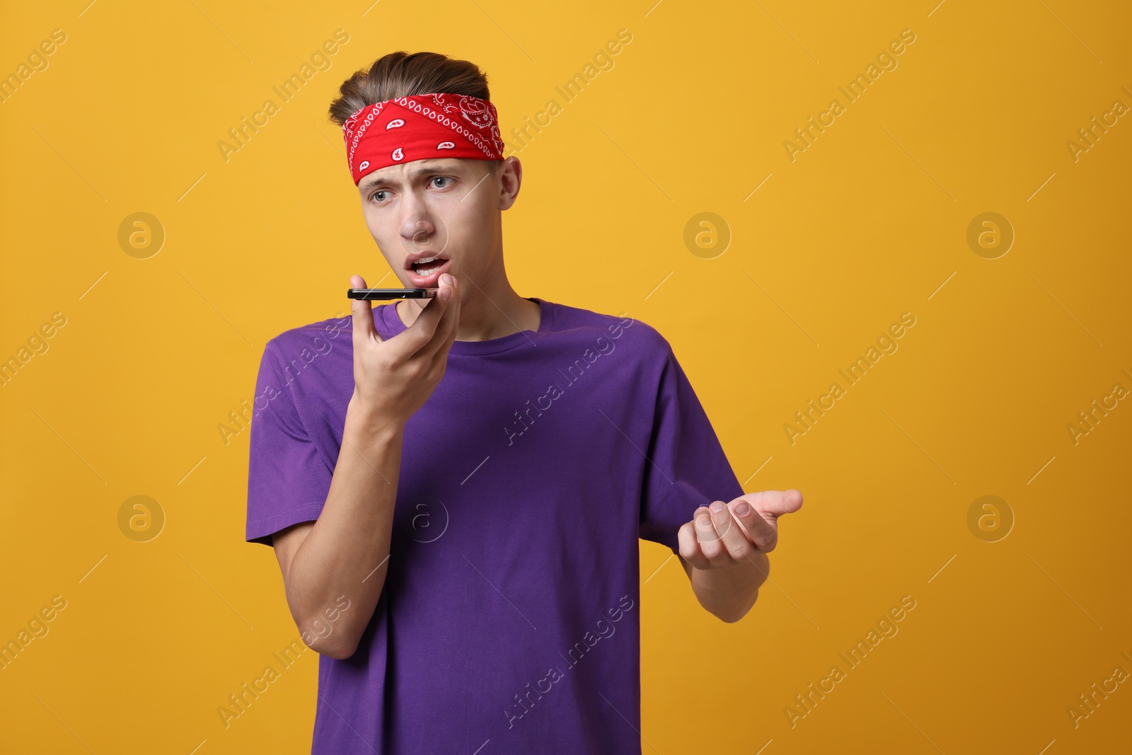 Photo of Young man recording voice message via smartphone on orange background