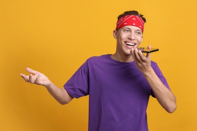 Young man recording voice message via smartphone on orange background