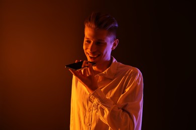 Young man recording voice message via smartphone on color background with neon lights