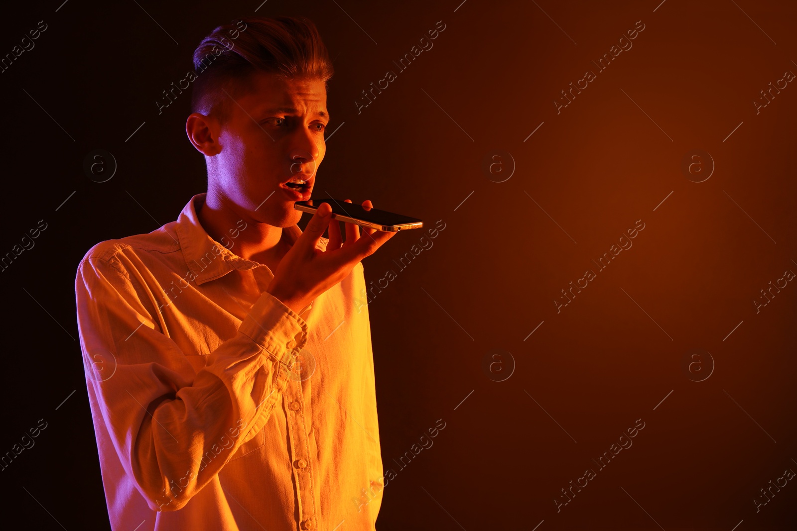 Photo of Young man recording voice message via smartphone on color background with neon lights, space for text