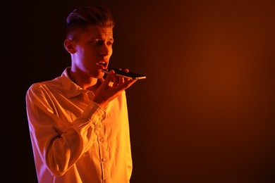 Young man recording voice message via smartphone on color background with neon lights, space for text