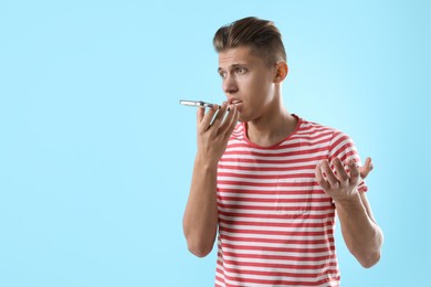 Young man recording voice message via smartphone on light blue background, space for text