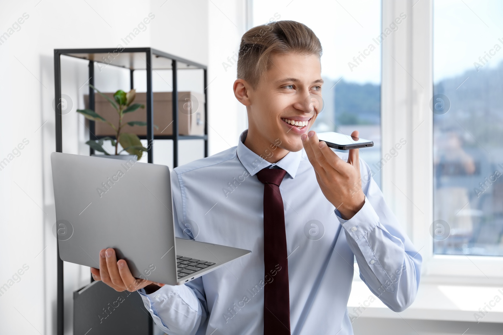 Photo of Young man with laptop recording voice message via smartphone in office