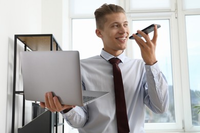 Young man with laptop recording voice message via smartphone in office