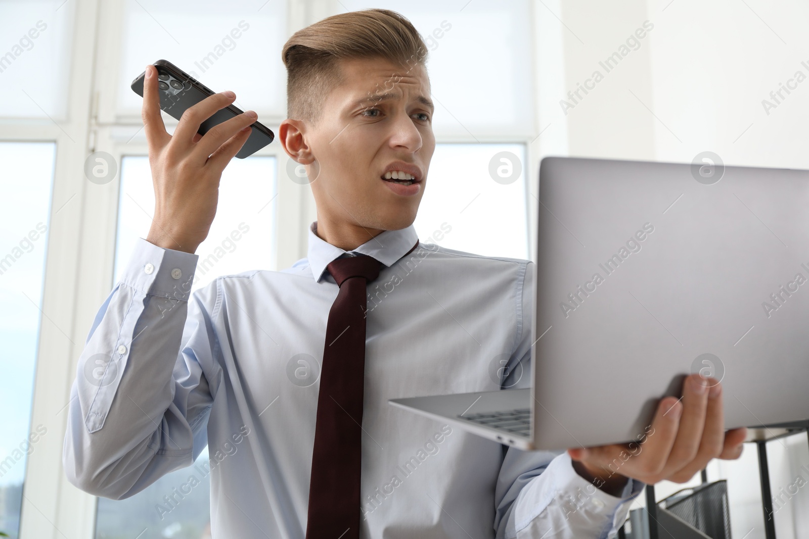 Photo of Young man with laptop listening to voice message via smartphone in office