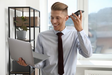 Photo of Young man with laptop listening to voice message via smartphone in office