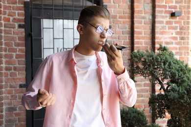 Photo of Young man recording voice message via smartphone outdoors