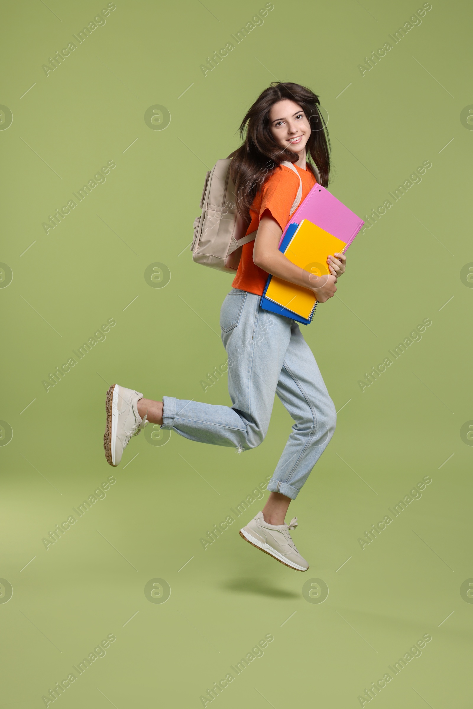 Photo of Cute teenage girl with backpack and books jumping on olive background