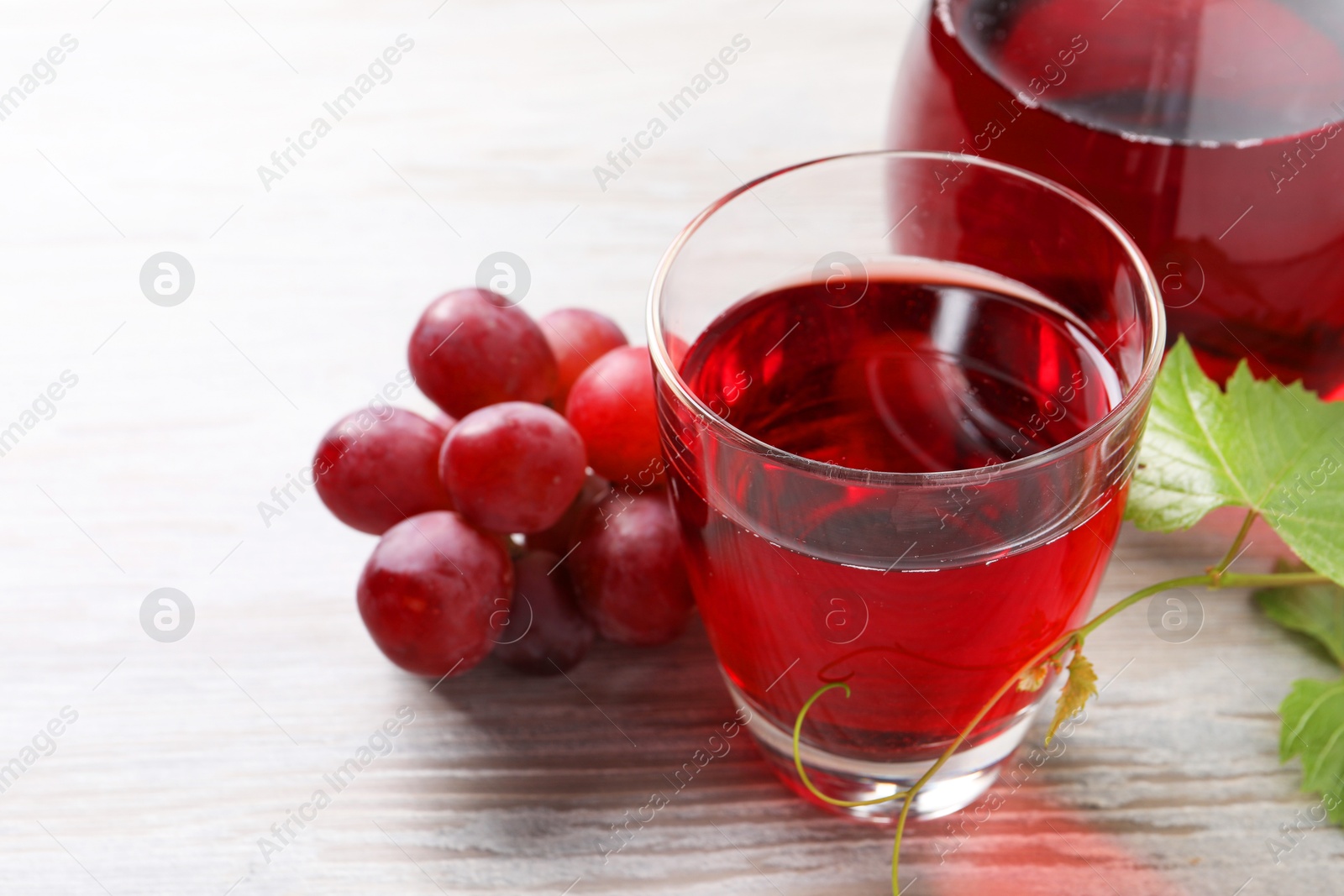 Photo of Tasty grape juice in glass, leaf and berries on light wooden table, closeup