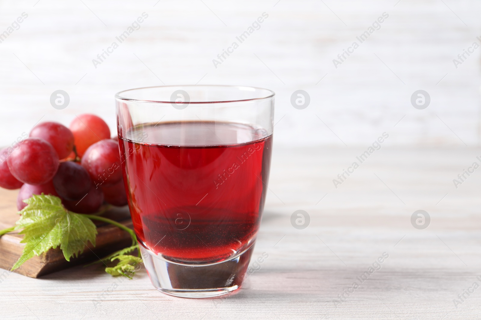 Photo of Tasty grape juice in glass, leaves and berries on light wooden table, closeup. Space for text