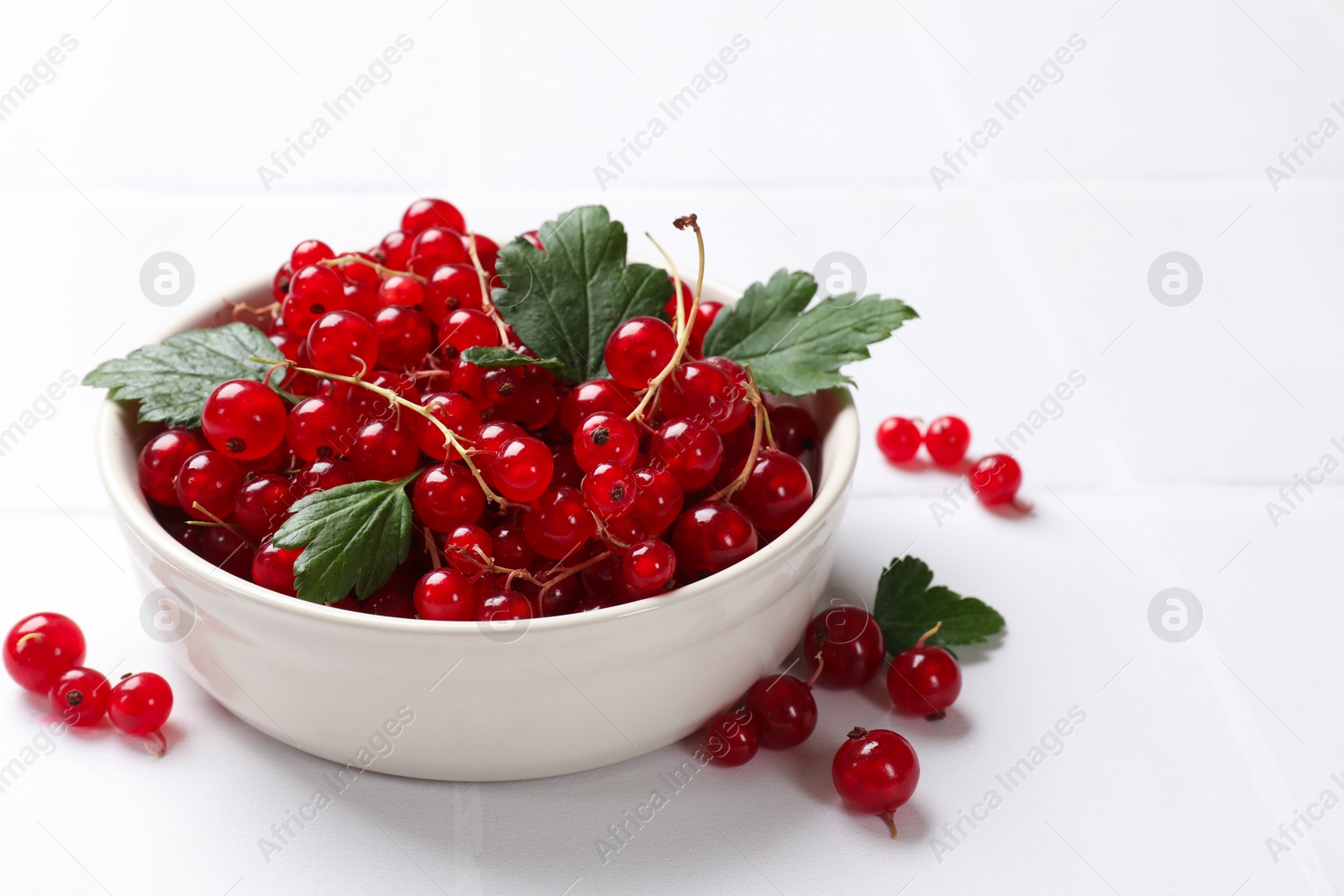 Photo of Fresh red currants and leaves in bowl on white table, closeup. Space for text