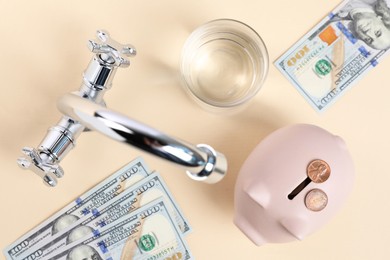 Photo of Water saving concept. Piggy bank, tap, glass of drink, dollar banknotes and coins on pale yellow background, flat lay