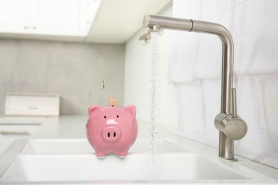 Photo of Water saving concept. Coins falling into piggy bank and stream of water flowing from tap in kitchen