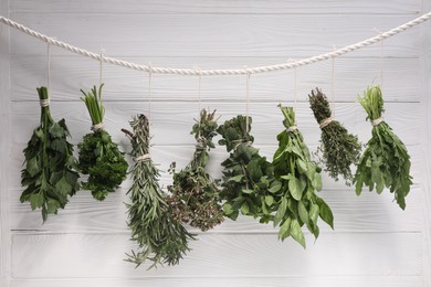 Photo of Bunches of different aromatic herbs hanging on rope near white wooden wall