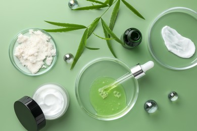 Photo of Petri dishes with cosmetic products, pipette and leaves on green background, flat lay