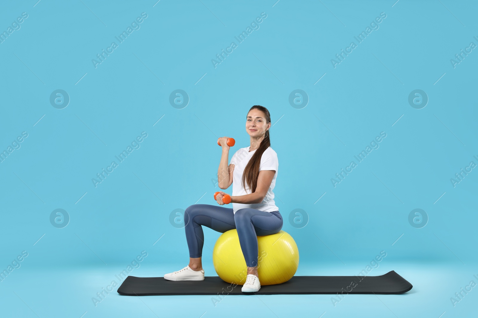 Photo of Beautiful pregnant woman with dumbbells doing exercises on fitball against light blue background