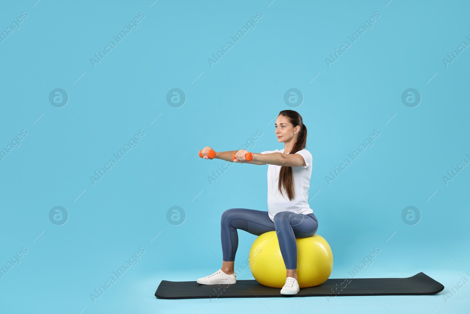 Photo of Beautiful pregnant woman with dumbbells doing exercises on fitball against light blue background