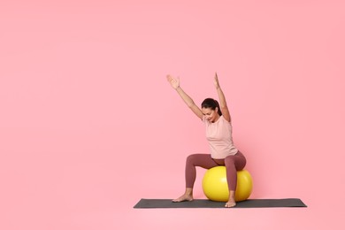 Photo of Beautiful pregnant woman doing exercises on fitball against pink background, space for text