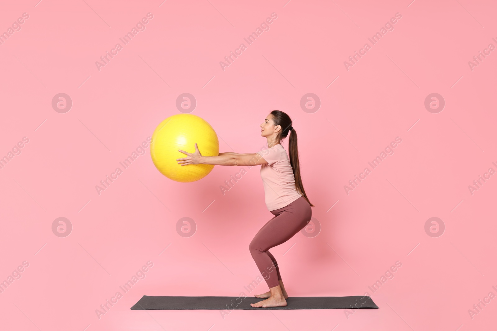 Photo of Beautiful pregnant woman with fitball doing exercises on mat against pink background