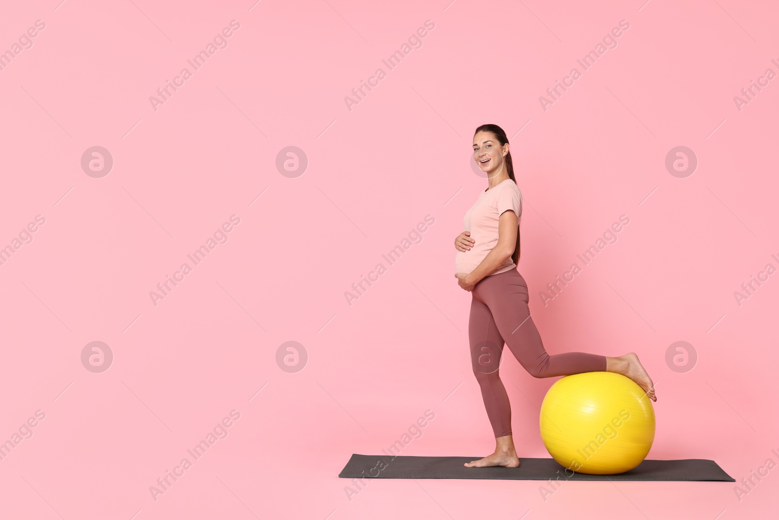 Photo of Beautiful pregnant woman with fitball doing exercises on mat against pink background, space for text