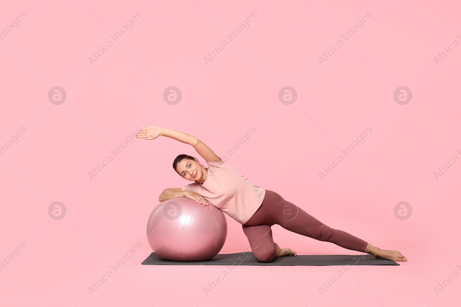 Photo of Beautiful pregnant woman with fitball doing exercises on mat against pink background