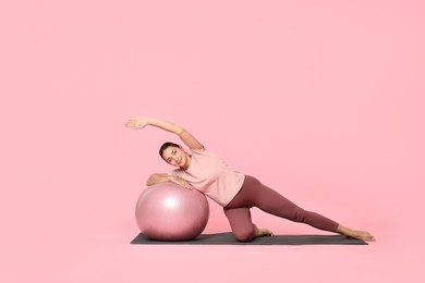 Beautiful pregnant woman with fitball doing exercises on mat against pink background