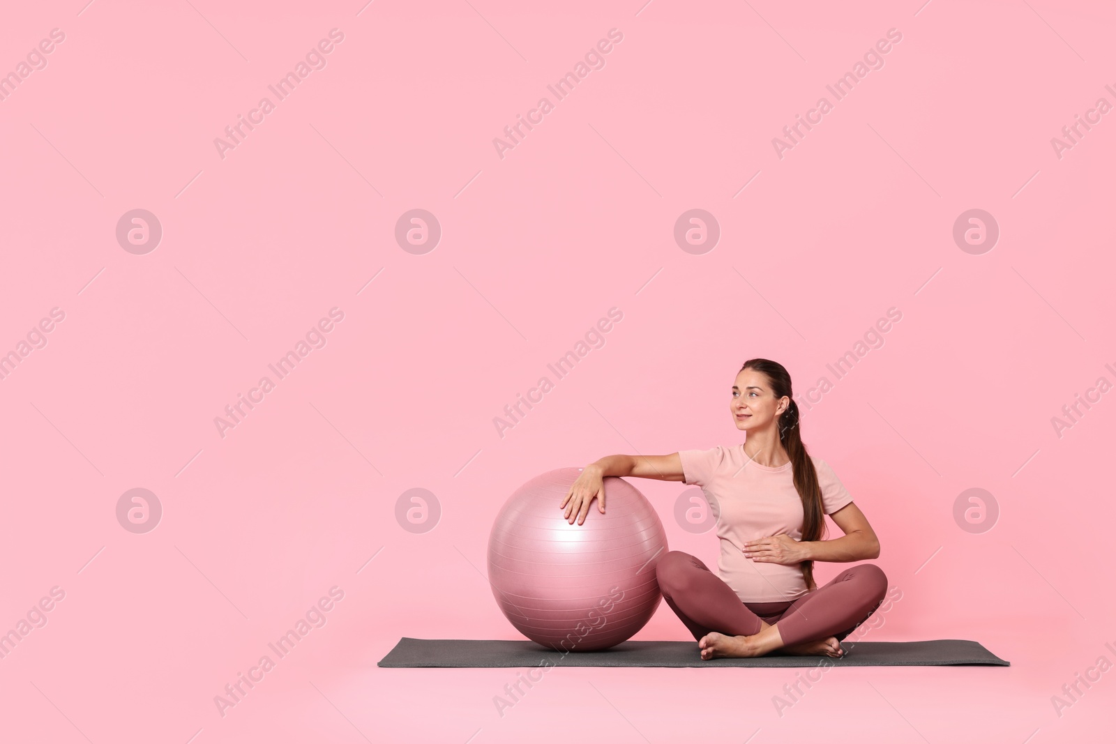 Photo of Beautiful pregnant woman with fitball on exercise mat against pink background, space for text