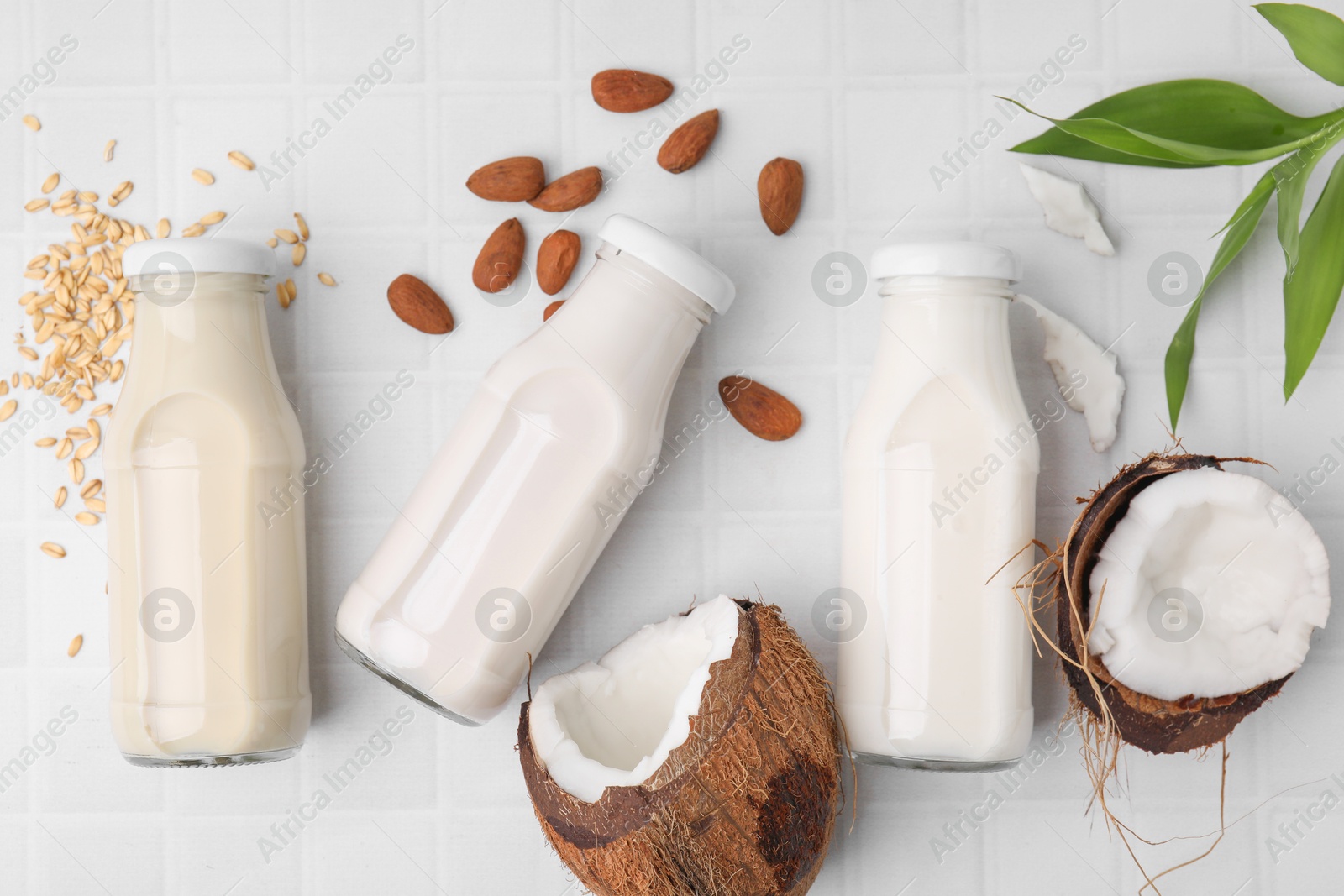 Photo of Different types of vegan milk in bottles with ingredients and green leaves on white tiled table, flat lay