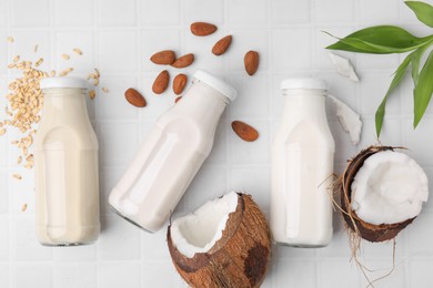 Photo of Different types of vegan milk in bottles with ingredients and green leaves on white tiled table, flat lay