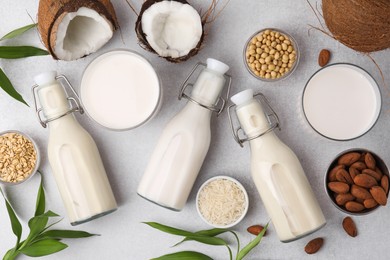 Different types of vegan milk with ingredients and leaves on light grey table, flat lay