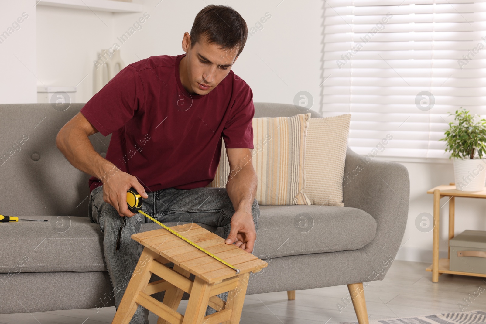 Photo of Man using tape measure while repairing wooden stool indoors